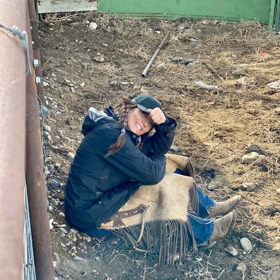 woman sitting on the ground of a stall outside with chaps on and smiling to the camera